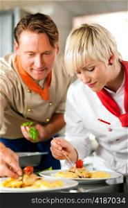 Two chefs in teamwork - man and woman - in a restaurant or hotel kitchen cooking delicious food, both are decorating the dishes