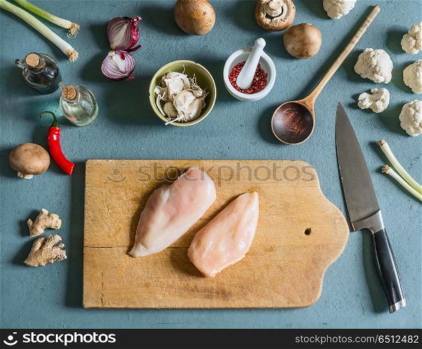 Two chicken breast on cutting board with knife on kitchen table background with ingredients , top view. Cooking preparation