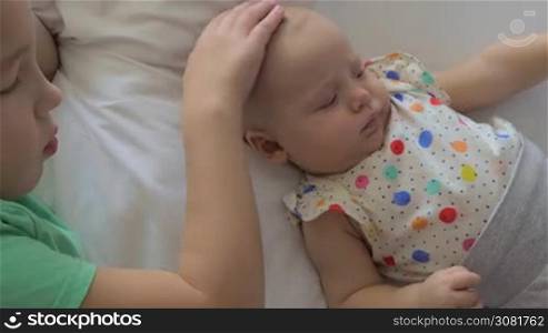 Two children are chilling on a white bed. A seven years old boy is taking care of his six months old baby sister. She is lying close to him, wearing a colorful polka dot shirt and looking a little bit grumpy. The boy is softly stroking on her head and gently blowing on it