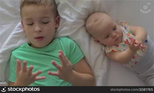 Two children are lying on white sheets close to each other. A young boy in a green T-shirt and a six months old baby girl in a colorful polka dot shirt. Both of them are looking at their hands. A boy is counting fingers and a baby girl just exploring her physicality