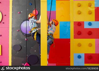 Two children in helmets climb on climbing wall in entertainment center, young climbers. Boy and girl having fun on ropes, kids spend the weekend on playground, happy childhood. Two children in helmets climb on climbing wall