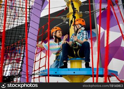 Two children in helmets climb on tightropes in entertainment center. Boy and girl having fun on ropes in climbing area, kids spend the weekend on playground, happy childhood. Two children in helmets climb on tightropes
