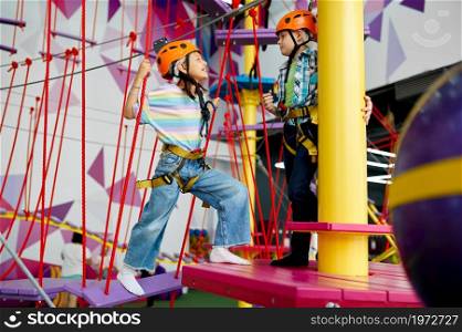 Two children in helmets climb on zip line in entertainment center. Boy and girl having fun on ropes in climbing area, kids spend the weekend on playground, happy childhood. Two children in helmets climb on zip line
