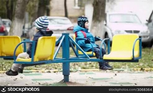 Two children on a merry-go-round sitting on the seats wrapped up warmly against the cold weather spinning around