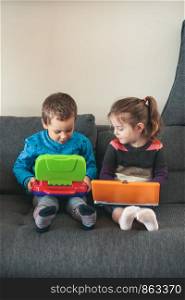 Two children playing with laptops learning basic digits, characters, sounds and images. Little girl and boy sitting on sofa and playing together at home. Candid people, real moments, authentic situations