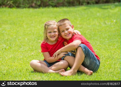 Two children who are brother and sister sit by each other with arms around the neck on the lawn as the smile and pose for a photograph.