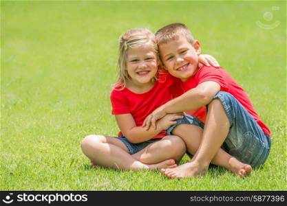 Two children whom are brother and sister are sitting on the green lawn with red t shirts and blue jean shorts with big smiles looking at the viewer at giving each other a big hug, on a beautiful summer day.