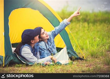 Two close friend Asian friendship relax in camping tent in green meadow on lake side view background. Girl pointing finger to sky. People lifestyle travel on vacation concept. Summer picnic activity