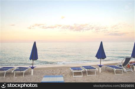 two closed umbrellas and some sun beds on a deserted beach with a calm sea at sunset