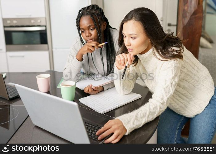 Two college girls studying together at home with laptops while drinking coffee. Multiethnic women.. Two college girls studying together at home with laptops while drinking coffee