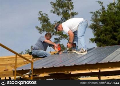 Two construction workers attach sheet metal to wood rafters of a barn with electric and battery powered screw guns.