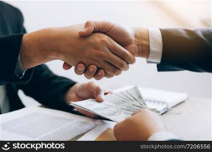 Two corporate businessmen shaking hands while one man places money on document in office room with corruption concept.