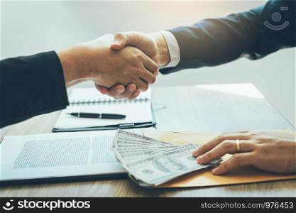 Two corporate businessmen shaking hands while one man places money on document in office room with corruption concept.
