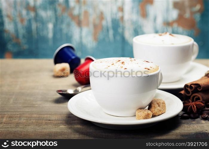 Two cups of cappuccino with coffee beans, spices and capsules on wooden background