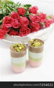 two cups with a puff dessert of a berry-chocolate-pistachio flavor, sprinkled with pistachios and chocolate. A bouquet of bright roses on the plates behind.