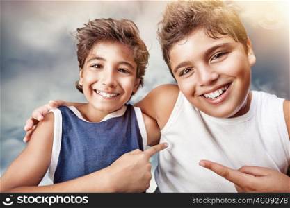 Two cute cheerful boys hugging and showing on each other by finger, portrait over cloudy sky background, happy children having fun at summertime