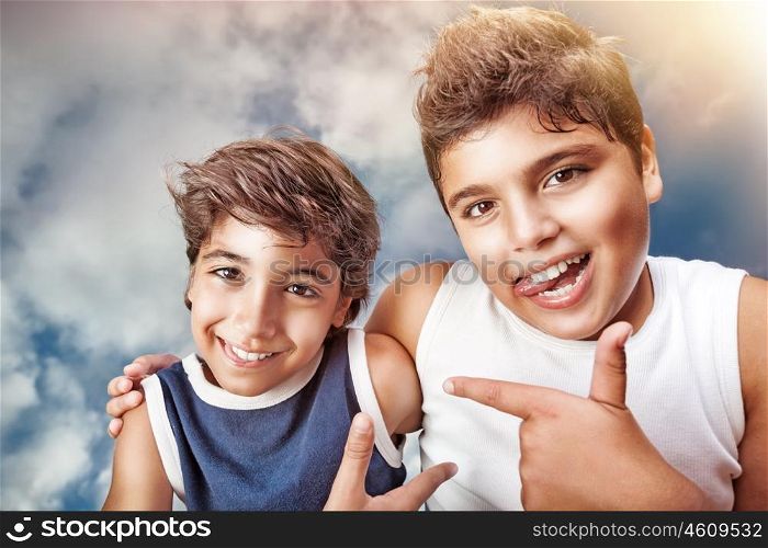 Two cute cheerful boys hugging and showing on each other by finger, portrait over cloudy sky background, happy children having fun
