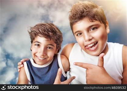 Two cute cheerful boys hugging and showing on each other by finger, portrait over cloudy sky background, happy children having fun