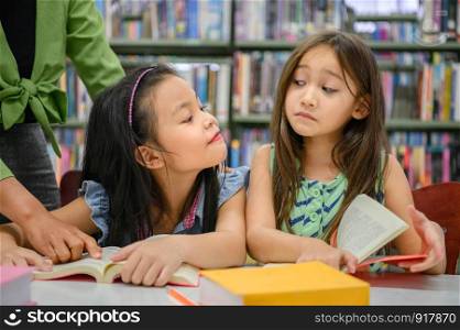 Two cute girls are jealous of each other while reading books in library while teacher teaching. People lifestyles and education. Young friendship and Kids relationship in school concept. Daycare theme