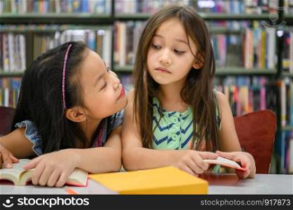 Two cute girls are jealous of each other while reading books in library while teacher teaching. People lifestyles and education. Young friendship and Kids relationship in school concept. Face to face