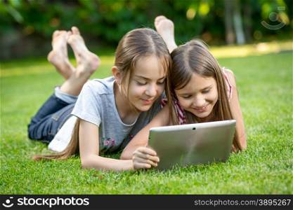 Two cute girls relaxing on grass at park and using digital tablet
