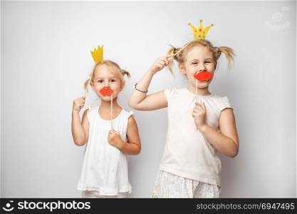 Two cute little girls with paper crown and red lips posing white background at home. Two cute little girls with paper crown and red lips posing white