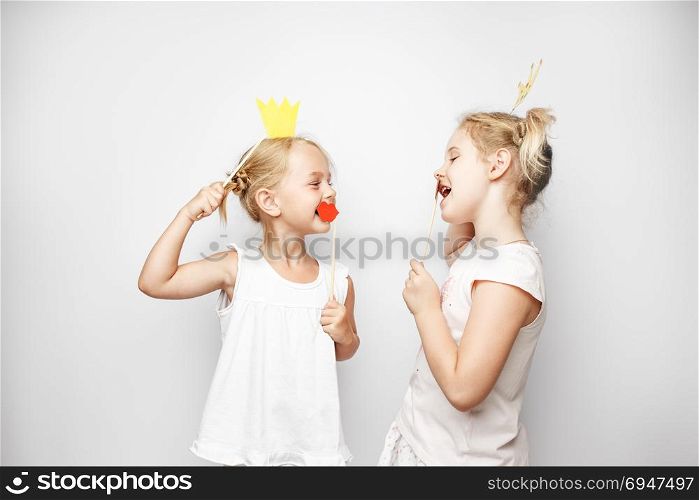 Two cute little girls with paper crown and red lips posing white background at home. Two cute little girls with paper crown and red lips posing white