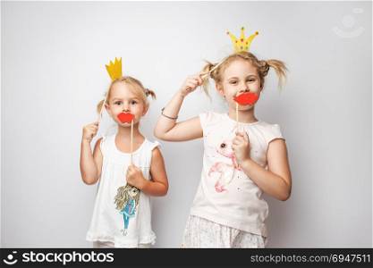 Two cute little girls with paper crown and red lips posing white background at home.. Two cute little girls with paper crown and red lips posing white background at home