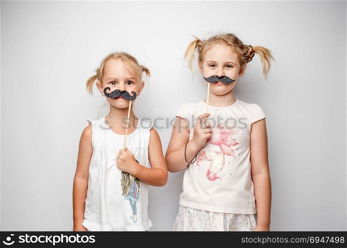 Two cute little girls with paper mustaches while posing against white background.. Two cute little girls with paper mustaches while posing against white background