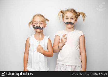 Two cute little girls with paper mustaches while posing against white background. Two cute little girls with paper mustaches while posing against