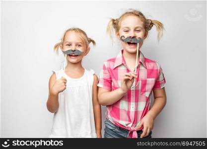 Two cute little girls with paper mustaches while posing against white background. Two cute little girls with paper mustaches while posing against