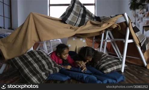 Two cute mixed race little girls watching funny video on digital tablet as they lie together in cubby house made of blanket and chairs. Smart kids using modern smart teghnology gadget in domestic room and smiling.
