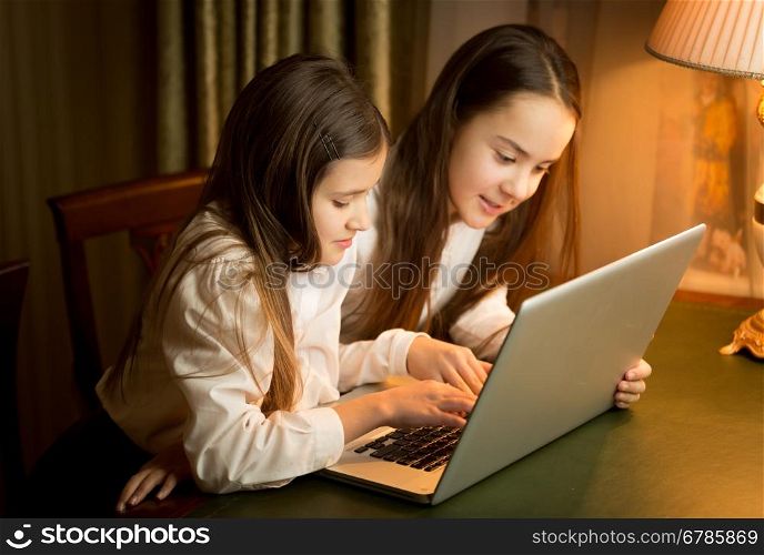 Two cute schoolgirls doing homework at laptop at night