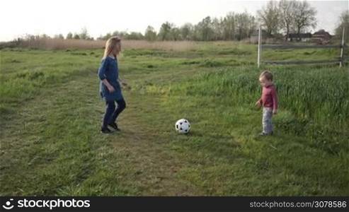 Two cute siblings having fun, playing soccer game in green lawn during spending leisure in countryside. Joyful toddler boy kicking a ball while his older beautiful sister teaching him playing football. Slow motion. Steadicam stabilized shot.