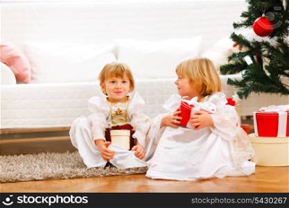 Two cute twins girl sitting with presents boxes near Christmas tree
