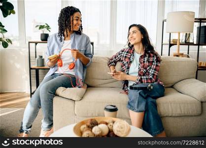 Two cute women enjoys listening to music on sofa with cup of coffe. Pretty girlfriends in earphones relax in the room, sound lovers resting on couch, female person using mobile phone. Two cute women enjoys listening to music on couch