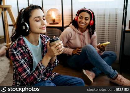 Two cute women enjoys listening to music with coffe at home. Pretty girlfriends in earphones relax in the room, sound lovers resting on couch, female friends leisures together. Women enjoys listening to music with coffe at home