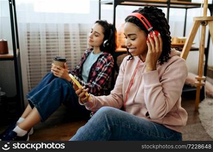 Two cute women enjoys listening to music with coffe at home. Pretty girlfriends in earphones relax in the room, sound lovers resting on couch, female friends leisures together. Women enjoys listening to music with coffe at home