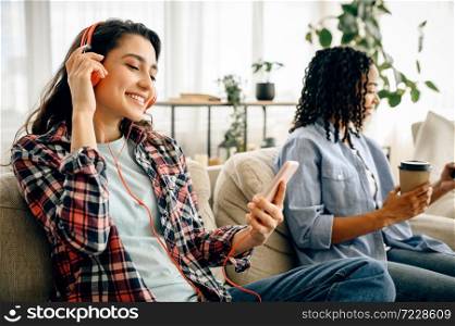 Two cute women in headphones leisures on sofa with cup of coffe. Pretty girlfriends in earphones relax in the room, sound lovers resting on couch, female person using mobile phone. Two cute women in headphones leisures on sofa