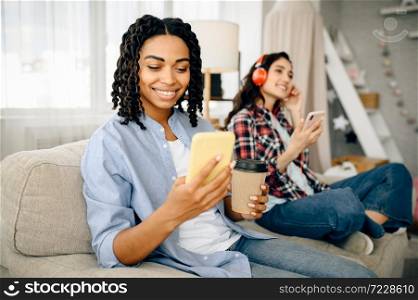 Two cute women in headphones listening to music and drinks coffe on sofa. Pretty girlfriends in earphones relax in the room, sound lovers resting on couch, female person using mobile phone. Women listening to music and drinks coffe on sofa