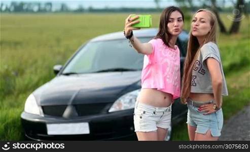 Two cute women stopped car on a roadside of rural road during trip. Young smiling women making selfies on the phone, posing, sending kisses and showing v sign.