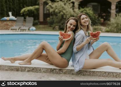 Two cute young women sitting on by the swimming pool and eating watermellon in the house backyard