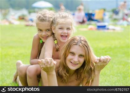 Two daughters are sitting on the mother's back, hot summer day relaxing on the green lawn beautiful beaches