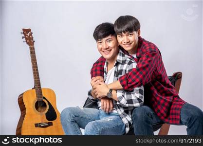 Two dear young men were sitting, cuddling in a chair, and with a guitar beside.