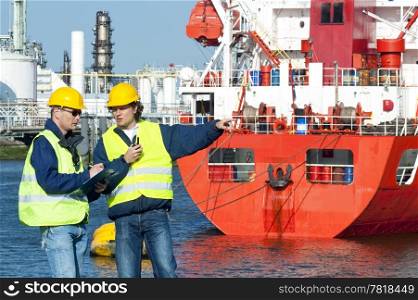 Two dockers at work, wearing safety vests hand hard hats, in frond of a red fire boat at a petrochemical harbor