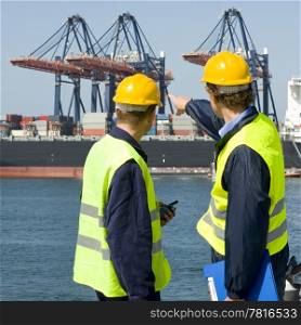 Two dockers in discussion, pointing at the unloading operations of a huge container ship