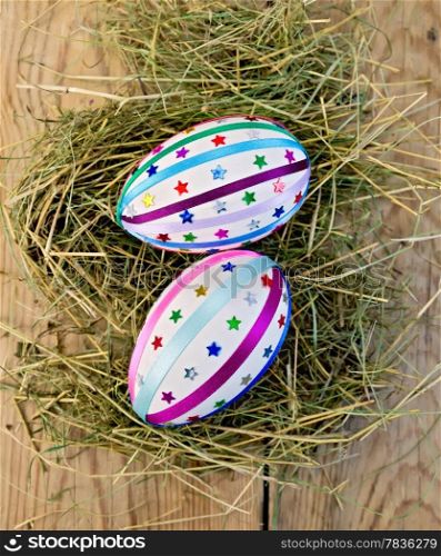 Two easter eggs, decorated with multicolored braid and sparkles as asterisks in the hay on the background of wooden boards on top