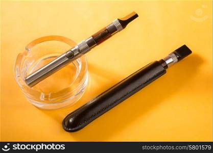 two electronic cigarettes, one in a clean glass ashtray, and the other in a black leather pouch, lie next to each other on a dark yellow background.
