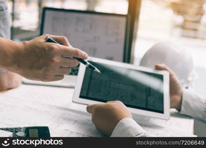 Two engineering working together and using digital tablet looking blueprint and analysis with architectural plan on desk.