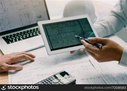 Two engineering working together and using digital tablet looking blueprint and analysis with architectural plan on desk.